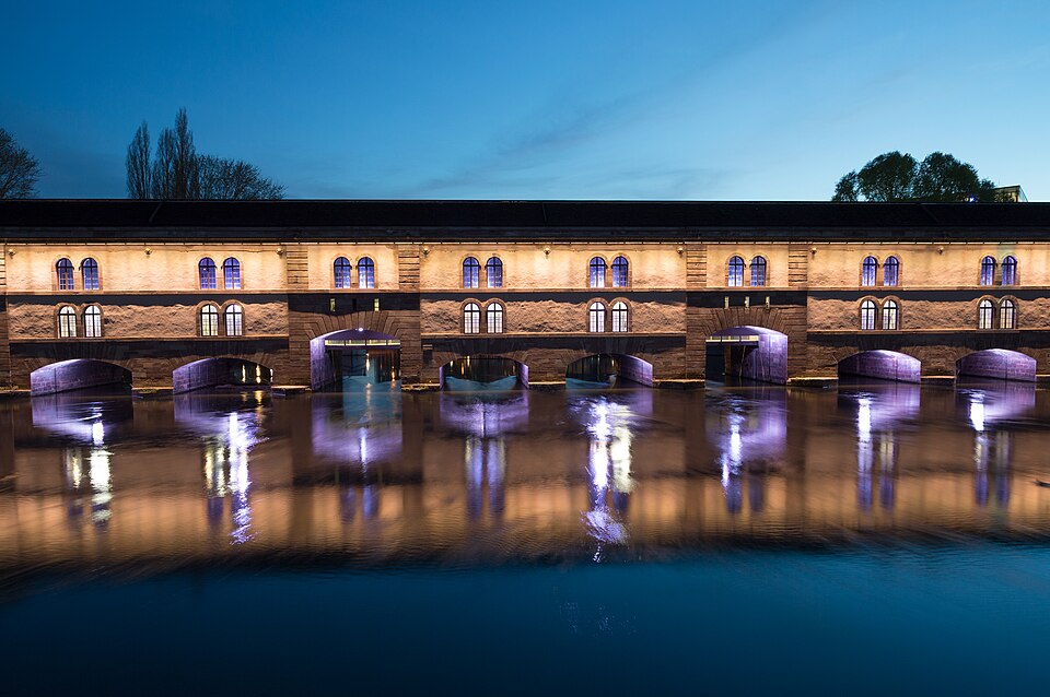The long sandstone arcade of the Barrage Vauban lit up at dusk, reflected in the Ill